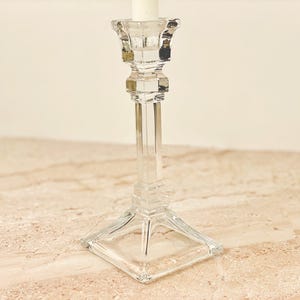 May include: A clear glass candlestick holder with a square base and a tall, slender stem. The candlestick is designed to hold a single candle. The glass has a faceted design, adding visual interest. The candlestick is on a light-colored surface.