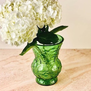 May include: A green glass vase with a textured surface holds a bouquet of white hydrangea flowers. The vase has a bulbous shape and sits on a light-colored surface.