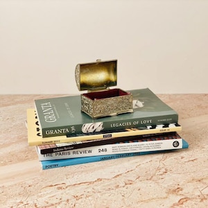 May include: An open, ornate, antique jewelry box with a red interior sits atop a stack of books. The books include "GRANTA," "LEGACIES OF LOVE," and "THE PARIS REVIEW 248." The books are stacked on a marble surface.