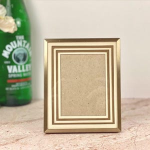 May include: A rectangular photo frame with a gold-colored border and a cream-colored inner border. The frame is empty, ready to display a photograph. A green glass bottle and a vase with white flowers are in the background.