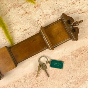 May include: A wooden key holder in a dark brown finish. The key holder has a rectangular design with decorative edges and a small, ornate top. A set of keys and a teal Michael Kors tag are attached.