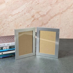 May include: A silver-colored, hinged photo frame with two rectangular openings. The frame is open, revealing a tan backing. A stack of books with titles like "The Paris Review" and "Granta" is to the left of the frame.