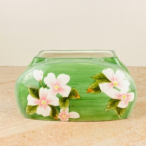 Puede incluir: Una jardinera de cerámica verde con un diseño floral pintado a mano. La jardinera presenta flores blancas y rosas con hojas verdes. La jardinera rectangular tiene lados redondeados y un acabado liso y brillante.
