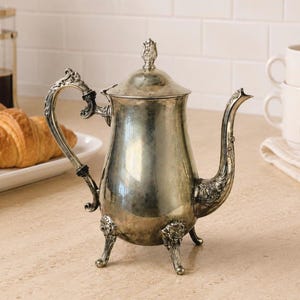 May include: A silver-toned metal teapot featuring an ornate, curved handle, a long spout with decorative detailing, and a lid topped with a small finial. The teapot stands on three short, sculpted legs and has a polished, reflective surface.