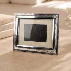 May include: A rectangular silver-toned picture frame with a polished metallic finish and a textured, grooved border design. The frame features a central rectangular opening with a light-colored matting and a dark, blank insert for a photograph.
