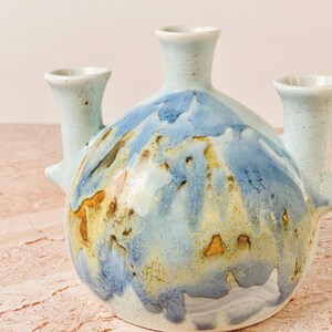 May include: A ceramic vase with three spouts and a rounded body. The vase is painted with a blend of blue, yellow, and brown hues, creating a unique, artistic design. The vase is a decorative item.