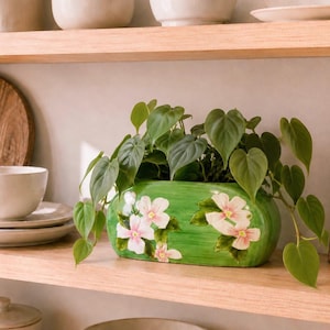 May include: An oval-shaped green ceramic planter featuring hand-painted white and pink dogwood flowers. A trailing heart-leaf philodendron plant with vibrant green foliage grows out of the top of the planter, which sits on a wooden shelf.