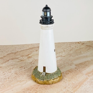 May include: A white lighthouse figurine with a black lantern room and gallery. The structure sits on a textured, green and tan base representing grassy terrain. The tower features a small rectangular doorway at the bottom and a single window.