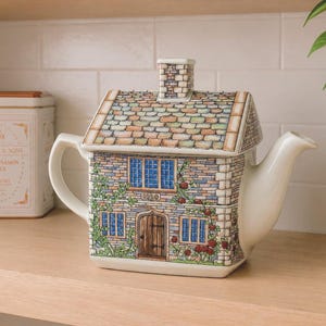 May include: A ceramic teapot shaped like a cottage with a stone-textured exterior, a tiled roof, and a chimney. The front features a wooden door, blue-paned windows, and climbing rose vines. The handle and spout are plain white ceramic.