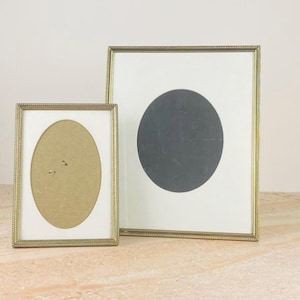 Peut inclure: Deux cadres dorés. L'un a une ouverture ovale avec un fond beige, l'autre un grand cercle noir sur fond blanc. Les deux cadres ont une bordure perlée et sont présentés sur une surface de couleur claire.