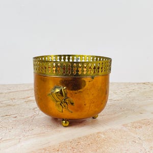 Puede incluir: Una maceta redonda de metal en tono cobre con un borde decorativo de color latón con un patrón de recortes repetitivo. El cuerpo exterior muestra un diseño floral en relieve y el recipiente se apoya sobre tres pequeñas patas metálicas redondeadas.