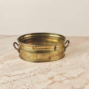 May include: A vintage brass planter with a round shape and two small handles. The planter has a slightly aged appearance, with visible wear and patina, suggesting it is an antique or a well-loved piece. The interior is visible.