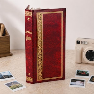 May include: A deep red photo album with a textured cover and gold foil detailing. The spine features the word "photos" in gold lettering. The album is rectangular with a vertical orientation and decorative gold borders on the front and spine.