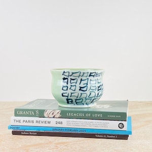 May include: A light green ceramic bowl with a dark blue geometric pattern sits atop a stack of books. The books have titles like "GRANTA" and "THE PARIS REVIEW". The bowl is a rounded shape, and the books are various sizes.