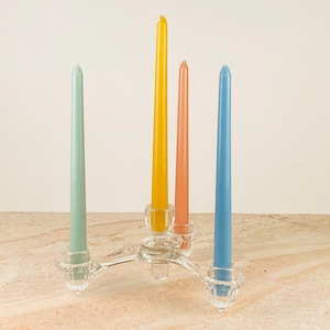May include: A clear glass candelabra holds four taper candles in pastel colors: light green, yellow, peach, and blue. The candelabra has three arms with decorative, scalloped edges. The candles are of equal height and stand on a light-colored surface.