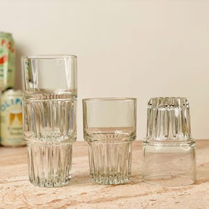May include: A collection of clear, stackable drinking glasses with a ribbed design. The glasses are arranged on a light-colored surface, with several cans of soda in the background. The glasses are ideal for serving beverages.