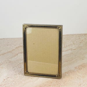 May include: A rectangular picture frame with a metallic gold finish and a beaded texture along the inner border. The frame features decorative corner accents and contains a plain, light brown cardboard insert behind the clear glass front.