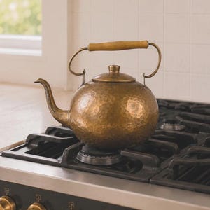 May include: A round, hammered brass tea kettle with a curved spout and a matching lid. The kettle features a horizontal wooden handle attached to a metal frame, resting on a black stovetop burner. The metal surface has a textured, dimpled finish.
