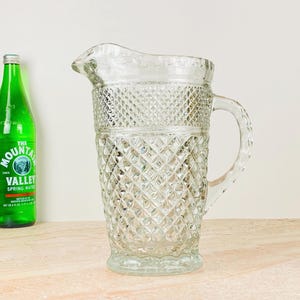 Peut inclure: Une carafe en verre transparent avec un motif en losanges, dotée d'une poignée et d'un bec verseur. La carafe est vide et repose sur une surface de couleur claire. Deux bouteilles de boissons sont visibles en arrière-plan.
