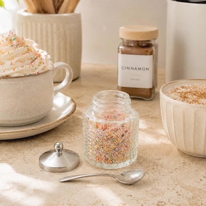 Könnte beinhalten: Ein Stillleben mit zwei Kaffeetassen, eine mit Schlagsahne und Streuseln, die andere mit Zimt. Ein Glas mit Streuseln, ein Glas mit Zimt und ein Kaffeebehälter sind ebenfalls vorhanden. Die Szene spielt sich auf einer hellen Oberfläche ab.