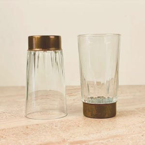 Può includere: Due bicchieri di vetro trasparente con fasce color rame. Un bicchiere è capovolto, l'altro in posizione verticale. Il vetro ha un design a coste verticali. Adatto per acqua o altre bevande.