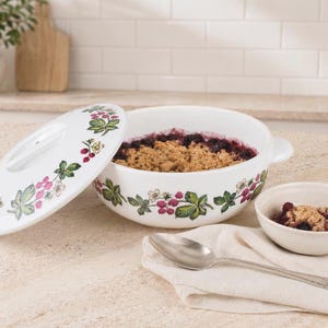 May include: A white ceramic casserole dish with a matching lid, decorated with a botanical pattern of blackberries, leaves, and flowers. The dish contains a fruit crumble, accompanied by a small matching bowl of crumble and a silver spoon.