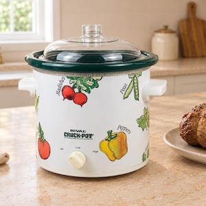 May include: A white Rival Crock-Pot stoneware slow cooker with a clear glass lid and a dark green rim. The exterior features colourful illustrations of vegetables including radishes, peas, and peppers with labels, and a single control knob for settings.