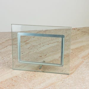 May include: A rectangular glass picture frame featuring a clear glass border and a central silver-toned metal frame insert. The frame is supported by a single silver-toned metal rod stand attached to the bottom center of the glass structure.