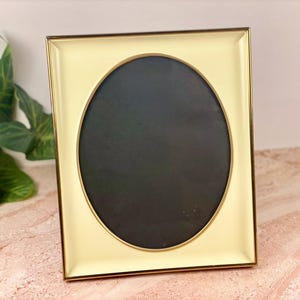 May include: A square, cream-colored picture frame with a gold trim and an oval opening. The frame is set against a pink and white surface with green leaves in the background. The frame is designed to hold a photograph or artwork.