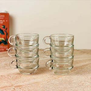 Può includere: Quattro tazze da tè in vetro trasparente impilate a coppie, ciascuna con un manico. Una scatola di tè Rishi Masala Chai è visibile sullo sfondo. Le tazze sono su una superficie marrone chiaro.