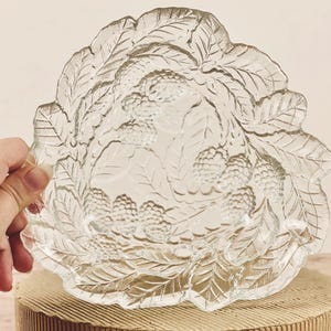 Puede incluir: Plato de servir de vidrio transparente con diseño de hojas y bayas. El plato tiene un borde festoneado y está sostenido por una mano. El diseño presenta hojas detalladas y racimos de bayas, creando un aspecto decorativo y elegante.