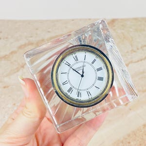 May include: A clear, faceted crystal desk clock with a square shape. The clock face is white with black Roman numerals and the text Waterford Crystal. It features a gold-toned circular bezel surrounding the dial and black clock hands.