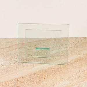 Peut inclure: Un cadre photo en verre transparent de forme rectangulaire. Le cadre présente un design en couches, avec un plus petit rectangle à l'intérieur d'un plus grand. Une petite pièce de verre rectangulaire est fixée au bas du cadre.