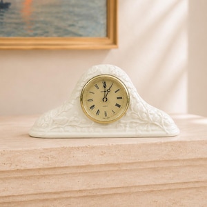 May include: A white ceramic mantel clock with a curved, embossed floral design and a round gold-toned bezel. The clock face is cream-colored with black Roman numerals and black hands, featuring the word Quartz printed below the center point.