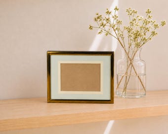 May include: A rectangular photo frame with a metallic gold finish, featuring a light mint green mat and a central beige insert. Beside the frame stands a clear glass vase containing a bouquet of small, delicate white dried flowers.