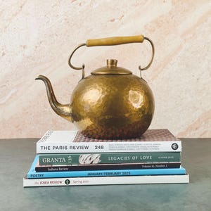 Op de afbeelding: Een vintage messing ketel met een houten handvat staat op een stapel boeken. De ketel heeft een gehamerde textuur en een gebogen tuit. De boeken zijn onder meer "The Paris Review", "Granta" en "The Iowa Review."