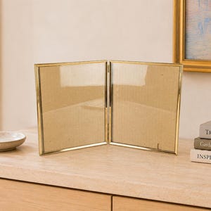 May include: A double-hinged picture frame with a thin, polished gold-tone metal border. The frame consists of two rectangular panels connected by a central hinge, allowing it to stand upright in a V-shape to display two separate photographs.