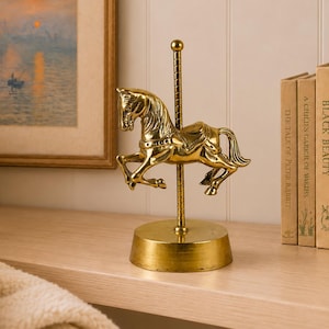 May include: A polished brass carousel horse figurine mounted on a central vertical pole with a circular base. The horse is depicted in a galloping pose with detailed mane, tail, and saddle features, standing as a standalone decorative object.