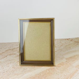 Peut inclure: Un cadre photo rectangulaire doté d'une bordure intérieure marron foncé et d'un contour extérieur fin en métal doré. Le cadre comprend une vitre transparente et un fond beige clair, conçu pour exposer des photos ou des œuvres en format vertical.