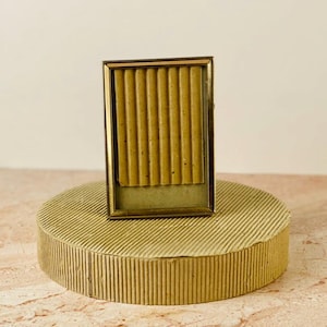 May include: A rectangular, gold-toned picture frame with a ribbed, textured interior. The frame is displayed on a round, gold-colored pedestal with a matching ribbed design. The background is a neutral beige.