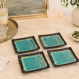May include: Four square ceramic appetizer plates. Each plate has a teal center with white linear designs and a dark brown border with gold accents. The plates are arranged on a light-colored surface.