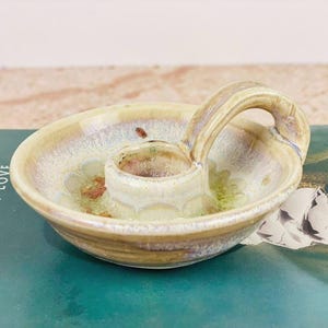 May include: A ceramic candle holder with a handle. The holder is a light beige color with brown and blue accents. The candle holder is sitting on a book with the words "Legacies of Love" on the side.