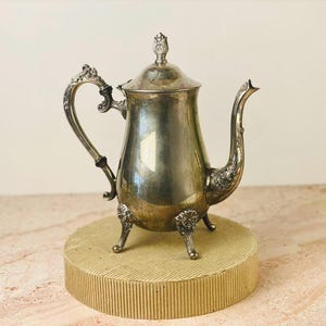 May include: A silver-colored antique teapot with ornate detailing on the handle, spout, and feet. The teapot has a lid with a decorative finial. It is displayed on a round, textured, beige platform.