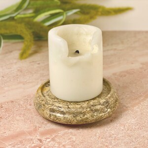 May include: A white pillar candle with a burnt wick sits on a decorative, round, stone coaster. The coaster has a mottled, tan and brown pattern. Green and yellow foliage is visible in the background.