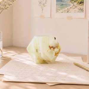 May include: A hexagonal paperweight crafted from pale green onyx stone with natural white and brown veining. The polished stone object sits on a piece of cream-colored stationery paper next to a gold-toned pen on a wooden surface.