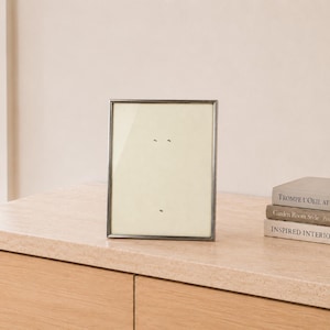 May include: A rectangular picture frame with a thin, dark metallic border and a cream-colored mat board. The frame is positioned upright, featuring a clear glass front and a neutral, light-colored interior display area with small, subtle markings.