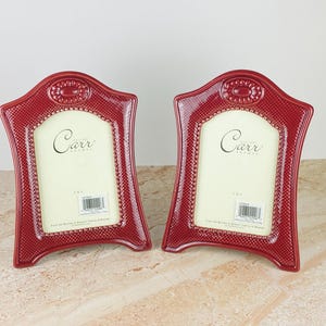 May include: Two matching red picture frames with a textured, dotted surface and a decorative oval emblem at the top center. Each frame features a cream-colored insert with the text Carr Frames and a barcode sticker on the lower portion.