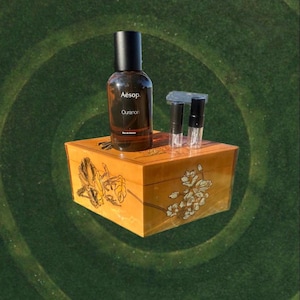 May include: A brown glass bottle of Aesop Ouranon Eau de Parfum with a black cap, resting on a decorative wooden box. The box is adorned with floral designs. Several small sample vials are also present.