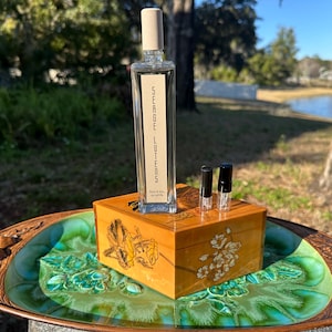 May include: A clear Serge Lutens perfume bottle with a light beige cap stands on a wooden box with floral designs. Two small black vials are next to the bottle. The box rests on a decorative, green-glazed ceramic tray.