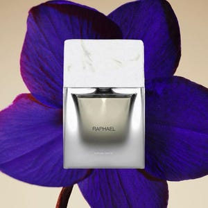 May include: A rectangular perfume bottle with a white marble-patterned cap and a silver metallic body. The front of the bottle features the text RAPHAEL in black lettering. The bottle is positioned in front of a large, vibrant purple orchid flower.